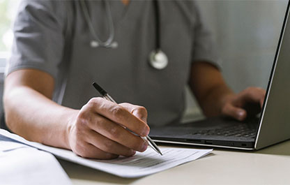 Doctor, face obscured, types on their computer with one hand and writes on an assessment form with the other.