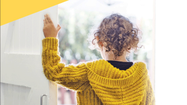 Child wearing a mustard knitted cardigan, looking beyond through the window of a full length door, holding the left arm against the frame.